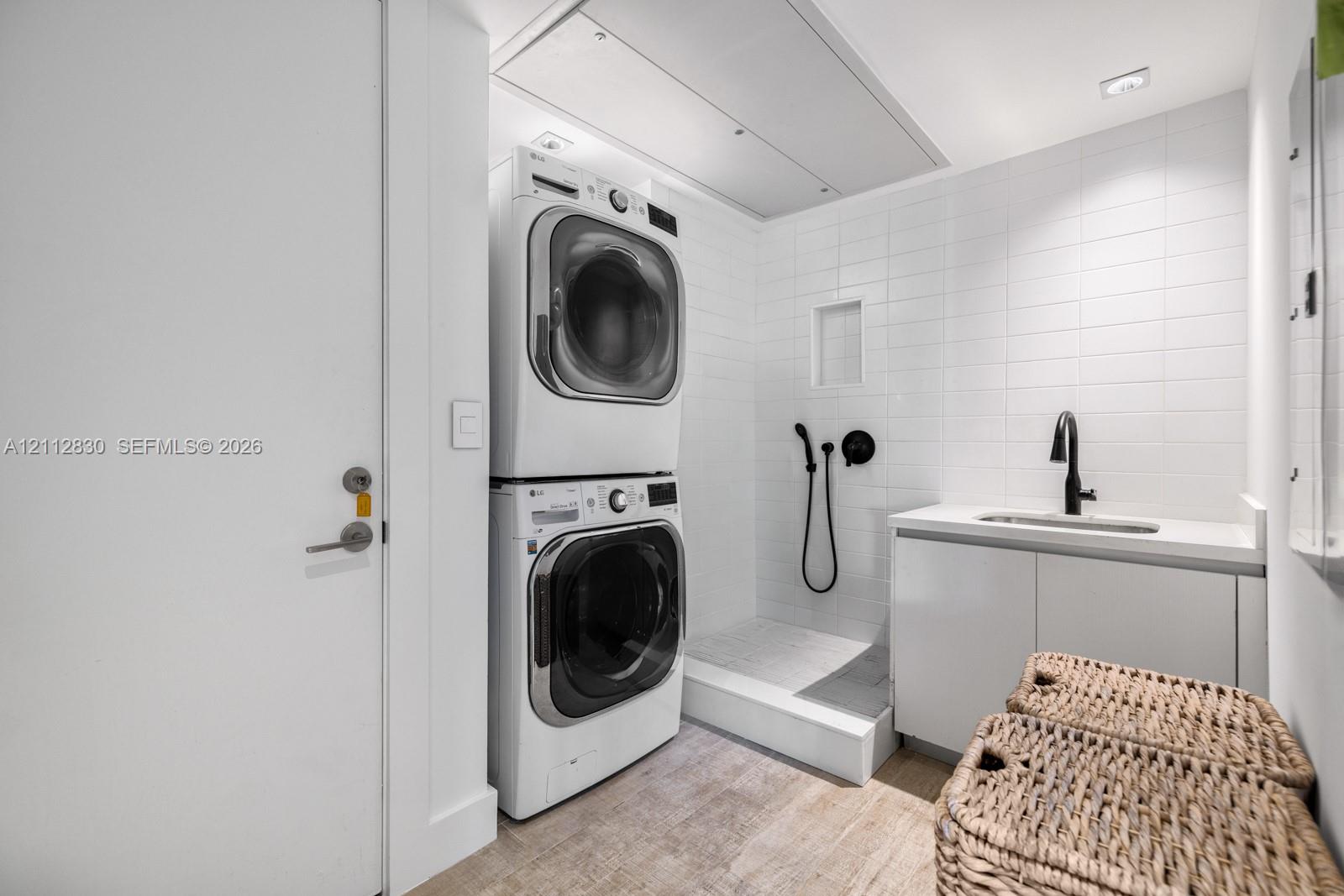 Laundry area featuring stacked appliances, tiled shower enclosure with handheld sprayer, built-in cabinetry, solid-surface countertop, and matte black faucet