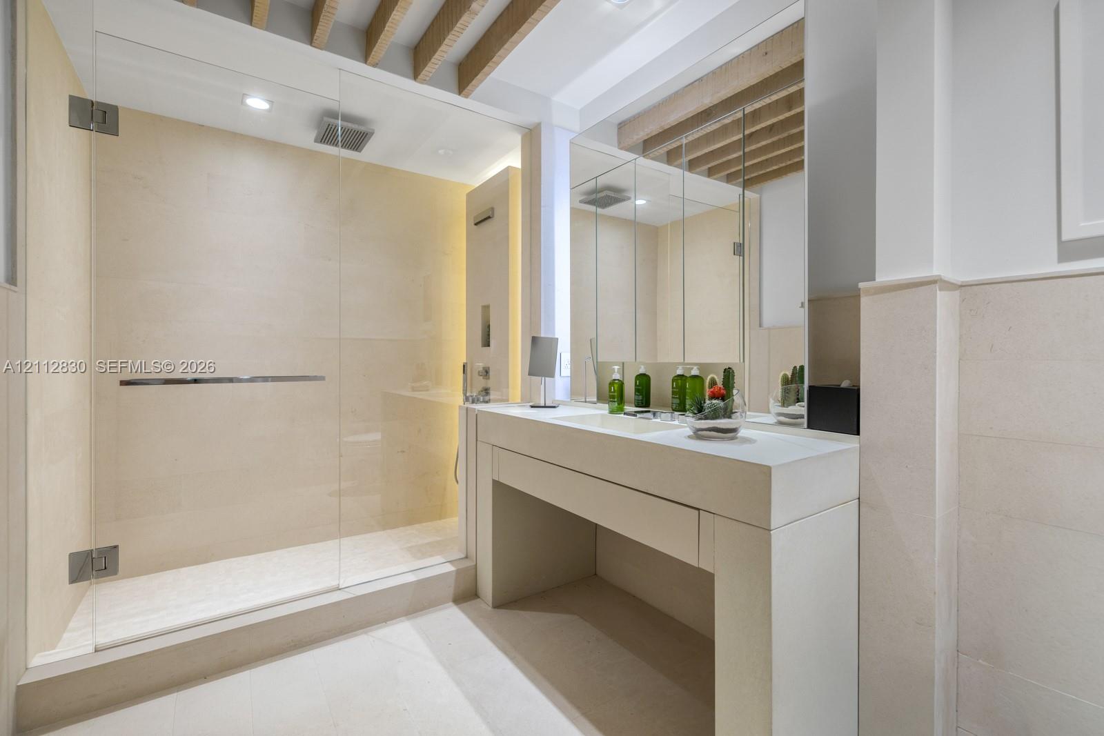 Stone-tiled shower featuring a frameless glass enclosure and rainfall showerhead, complemented by a custom stone vanity with an integrated sink and recessed ceiling beams