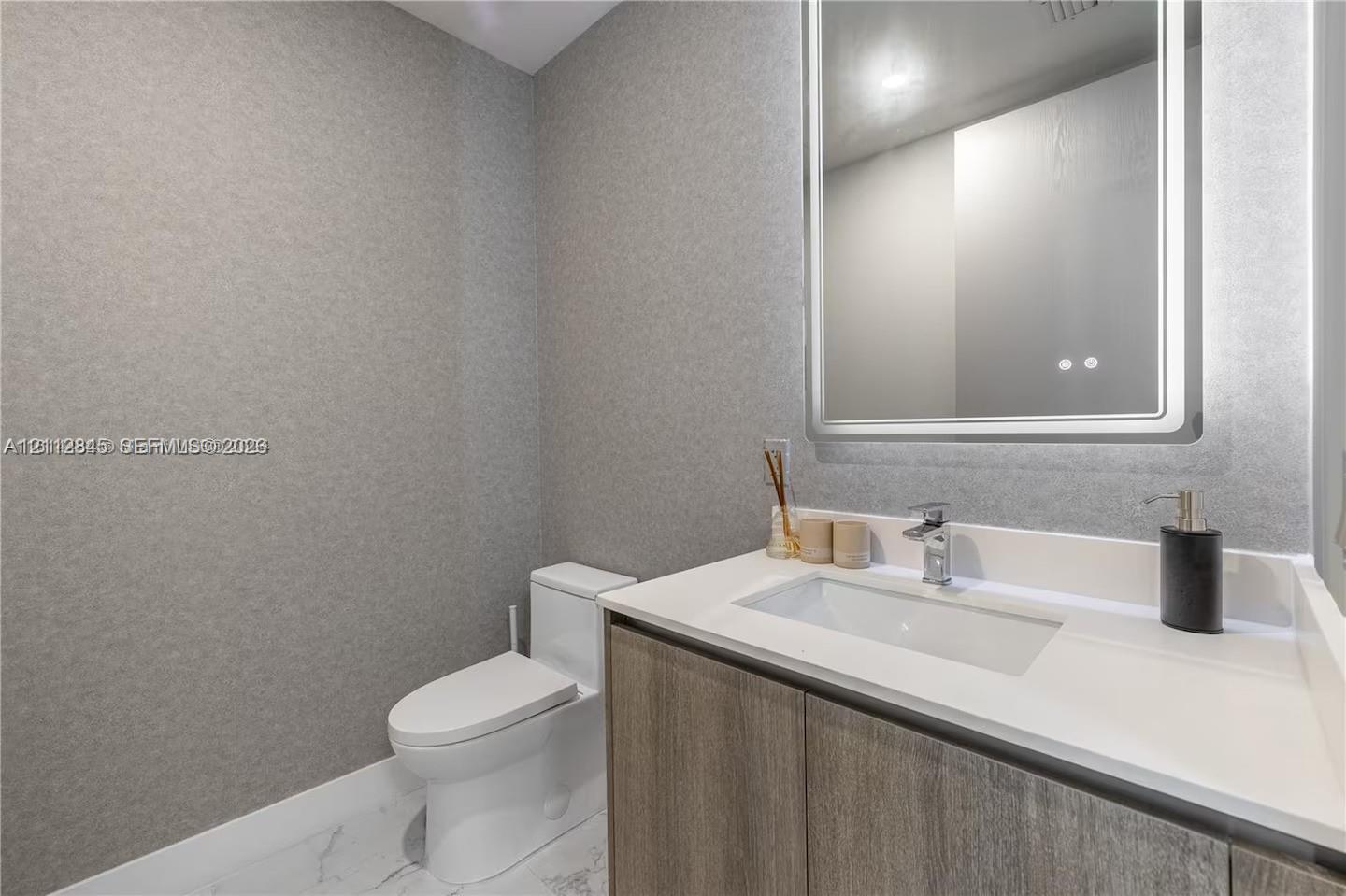Modern vanity with integrated rectangular sink and quartz countertop, complemented by a lighted rectangular mirror, textured wall covering, and stone-finish tile flooring