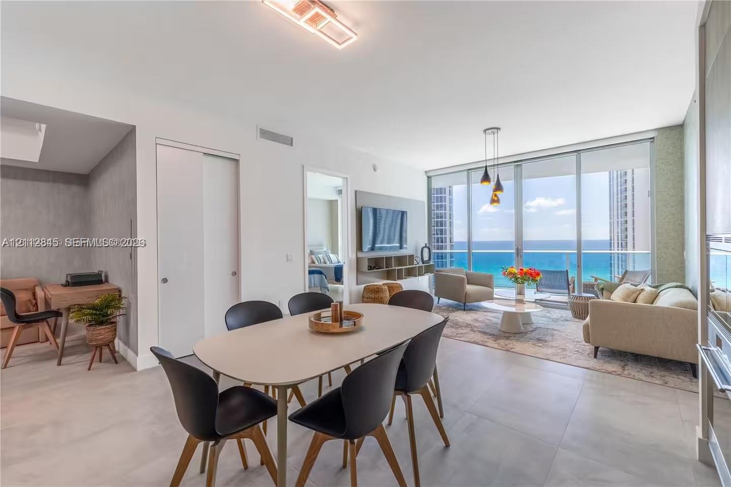 Open-concept interior featuring large-format tile flooring, floor-to-ceiling glass walls, and a balcony with an ocean backdrop Open-concept interior featuring large-format tile flooring, floor-to-ceiling glass walls, and a balcony with an ocean backdrop