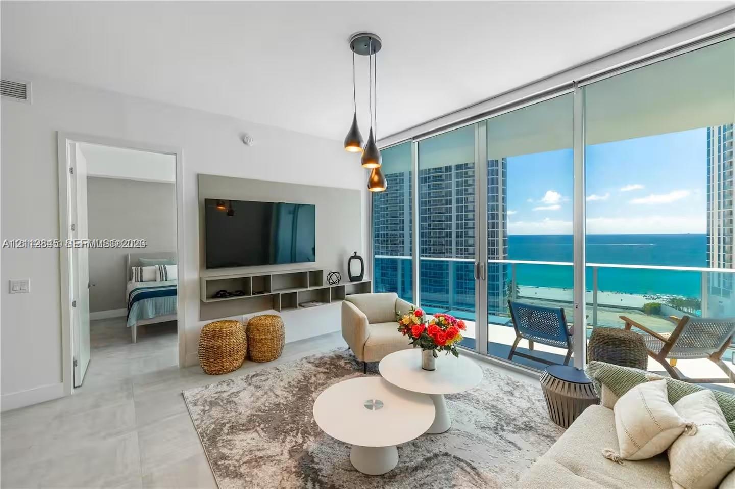 Floor-to-ceiling glass wall with ocean backdrop, private balcony, neutral large-format tile flooring, built-in wall-mounted media console, and triple-pendant light fixture Floor-to-ceiling glass wall with ocean backdrop, private balcony, neutral large-format tile flooring, built-in wall-mounted media console, and triple-pendant light fixture
