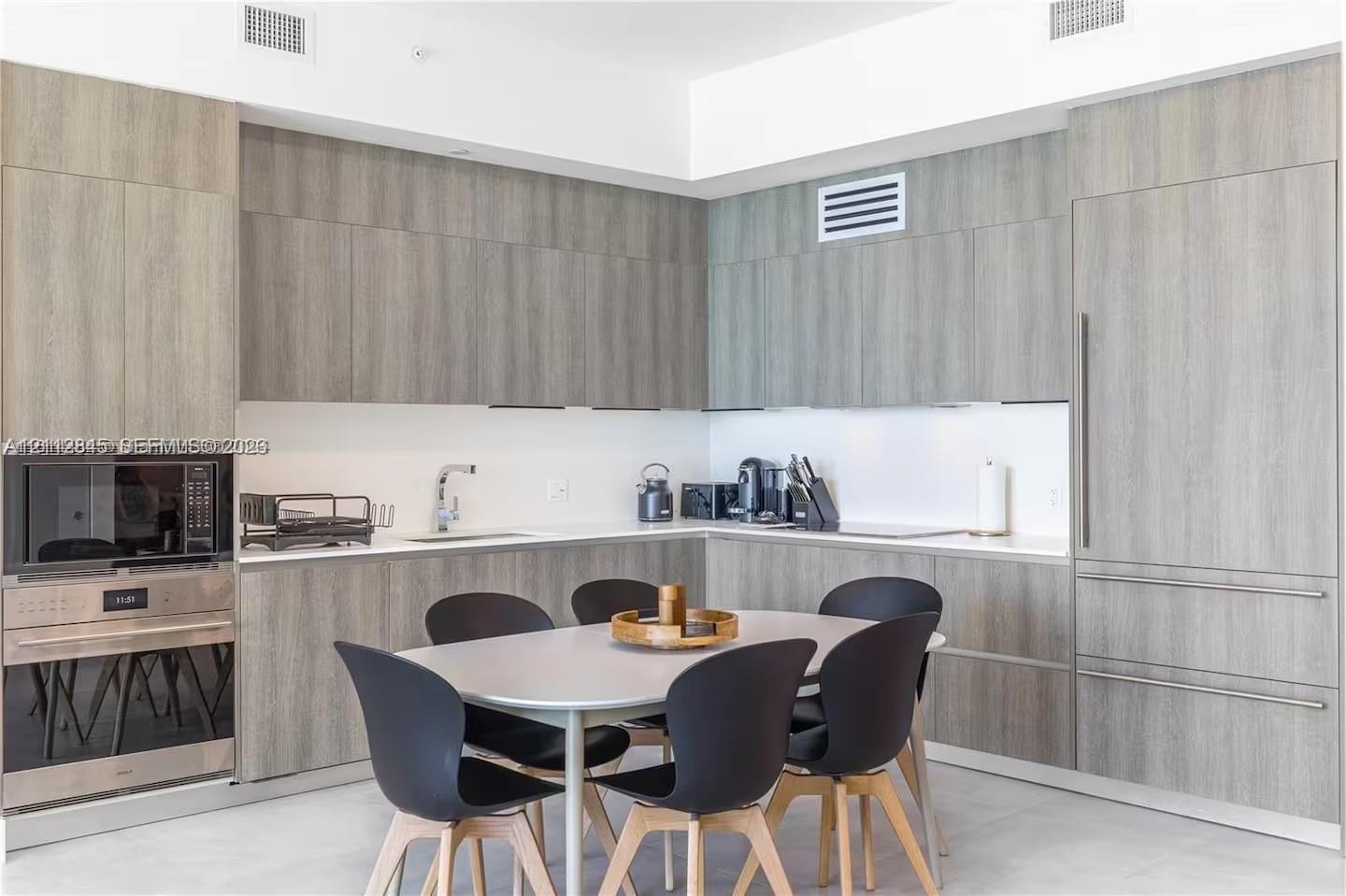 Kitchen featuring flat-panel wood-finish cabinetry, integrated refrigerator, built-in stainless steel microwave and wall oven, and light-toned solid surface countertops