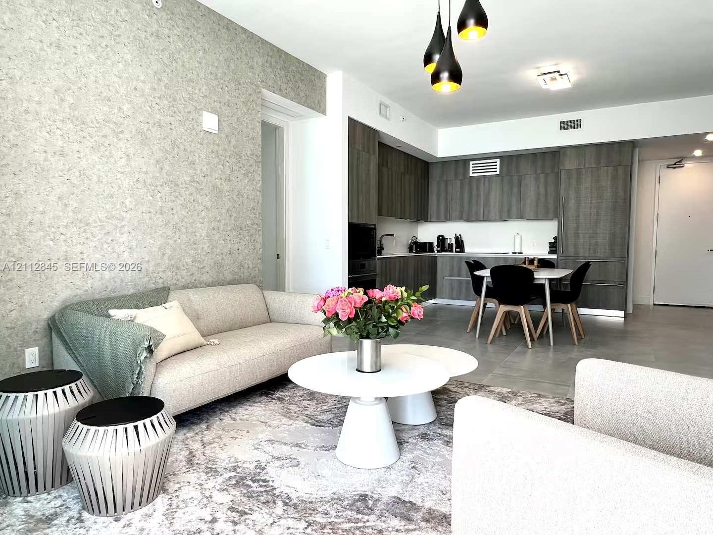 Open-concept living space featuring textured accent wallpaper, flat-panel dark wood-finish cabinetry, built-in stainless steel appliances, large-format tile flooring, and multi-pendant contemporary lighting