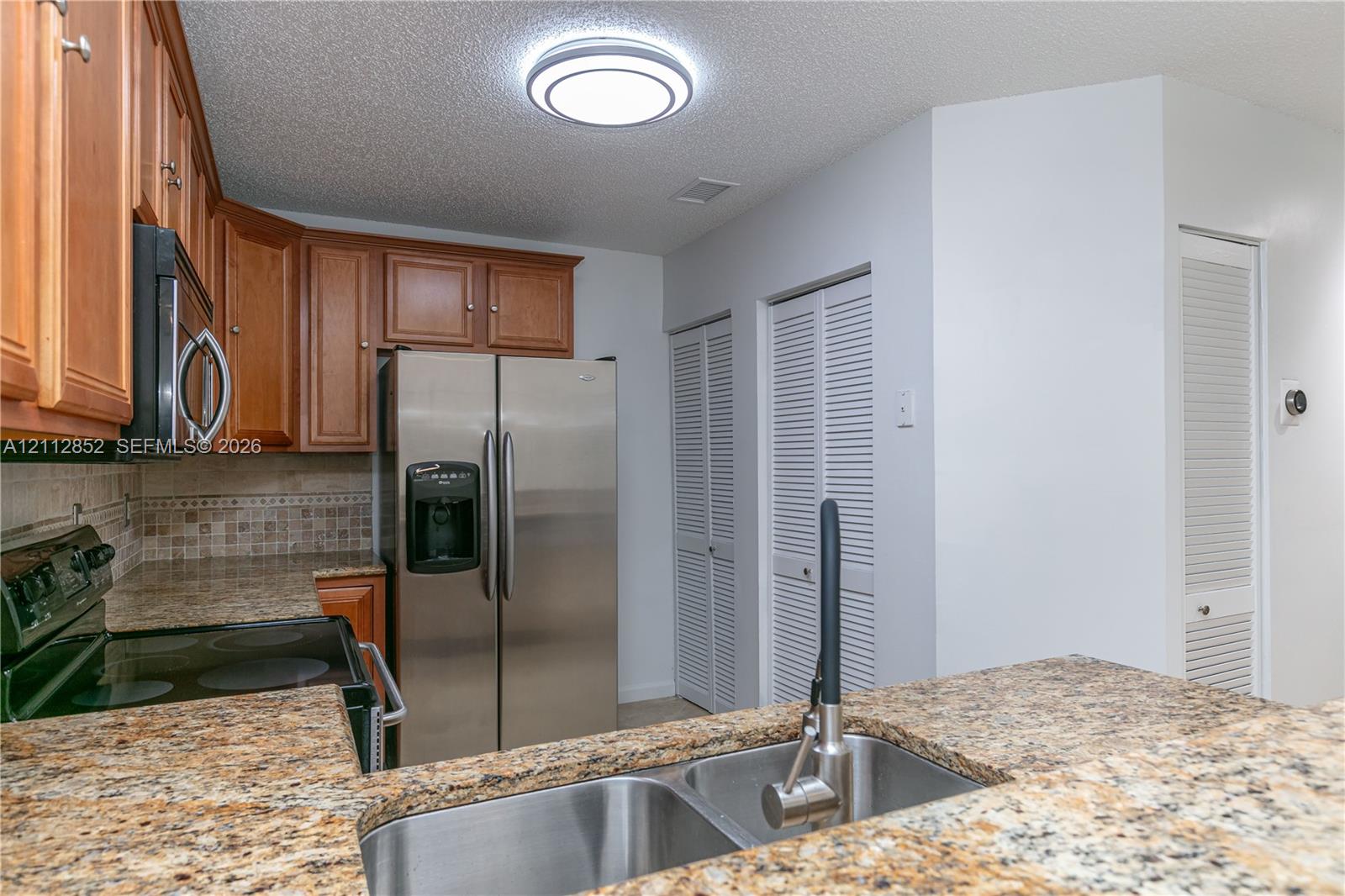 Kitchen featuring stone-patterned countertops, wood cabinetry, stainless steel appliances, dual-basin stainless steel sink with high-arc faucet, and tiled backsplash