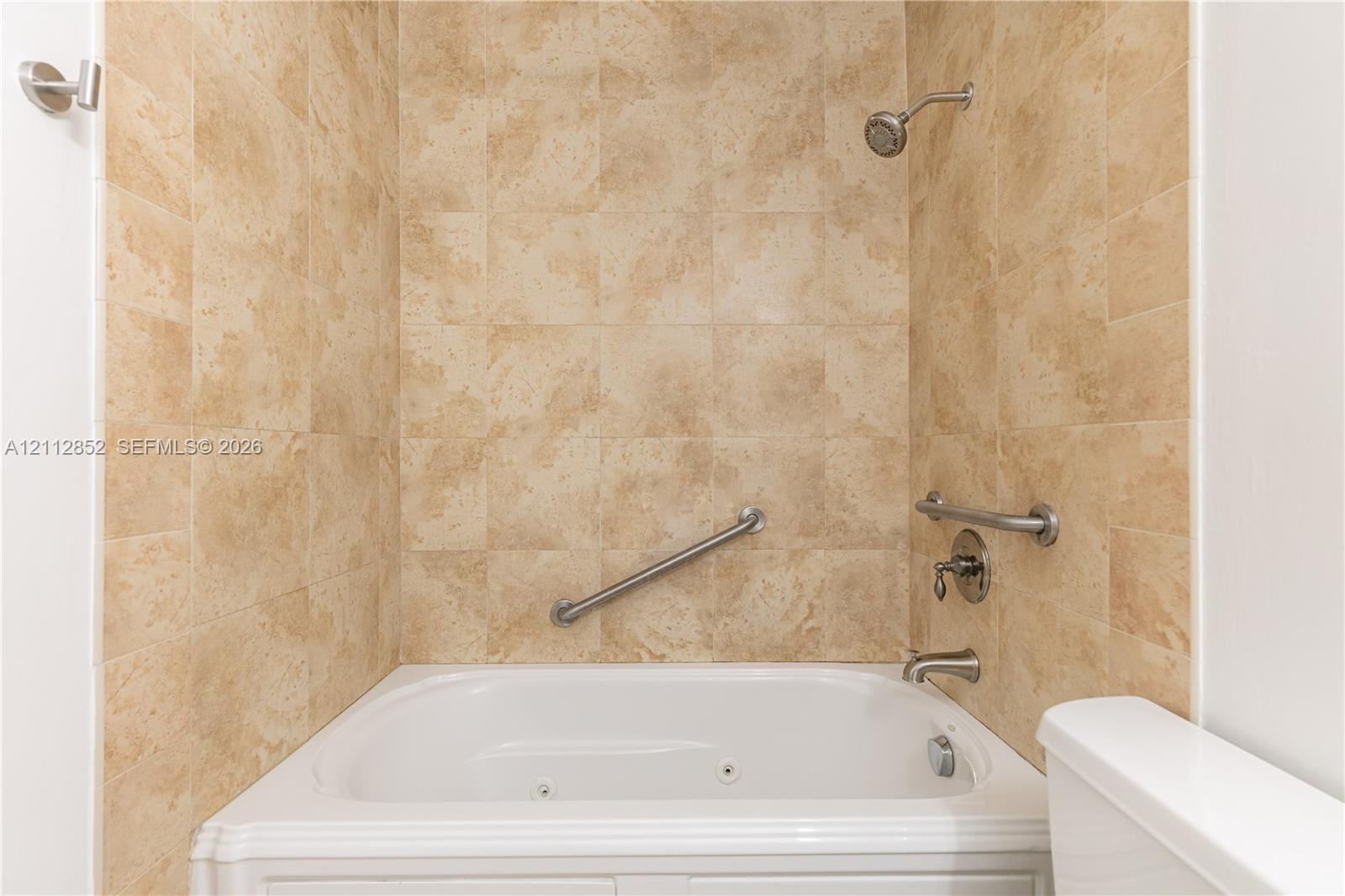 Jetted tub featuring stone-finish wall tiling, stainless steel wall-mounted grab bars, and a chrome shower fixture