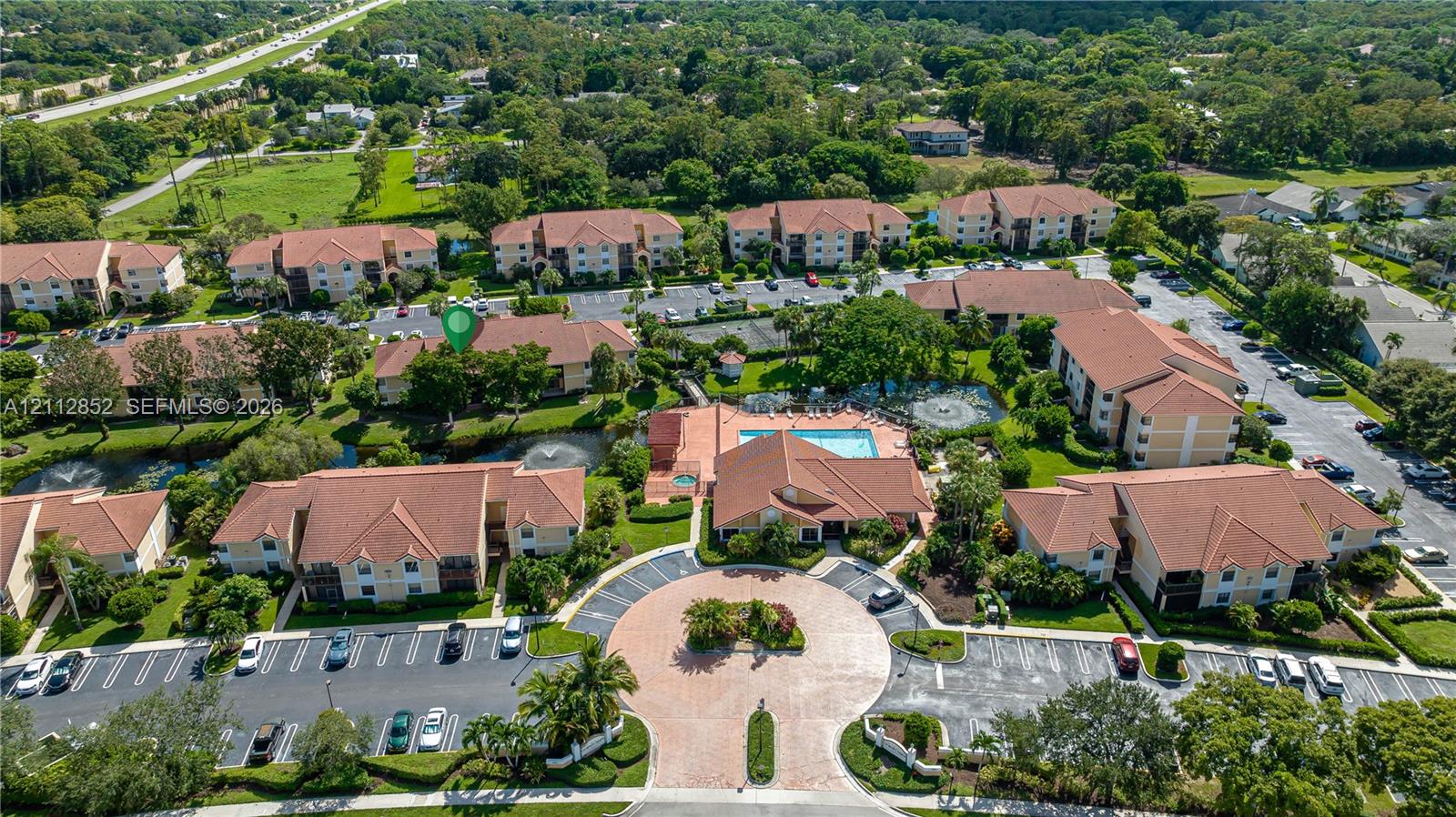 Aerial perspective of a multi-building residential complex featuring a central community swimming pool, paved roundabout entrance, designated parking areas, and landscaped grounds with water features