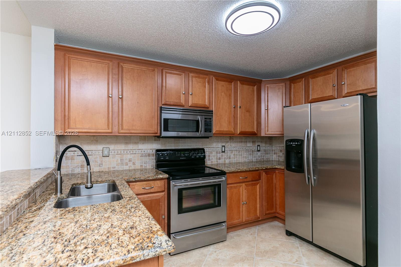 Kitchen featuring medium-tone wood cabinetry, stone countertops, stainless steel appliances, a tile backsplash, and a dual-basin undermount sink with a high-arch faucet