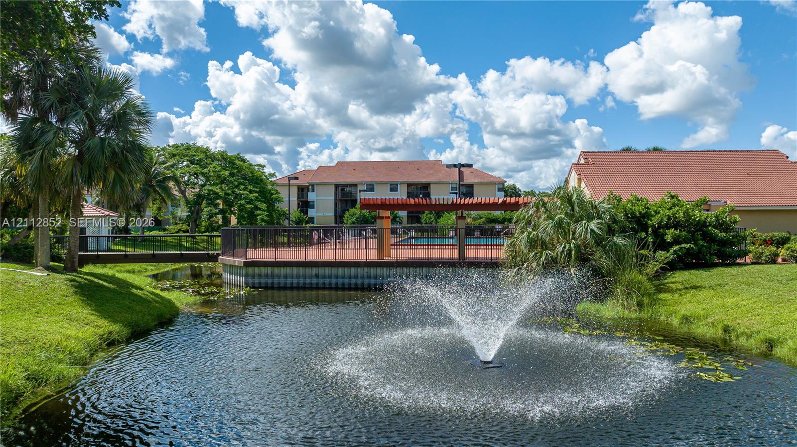 Community pond featuring a central fountain, manicured landscaping, pedestrian bridge, and paved pool deck with a wooden pergola