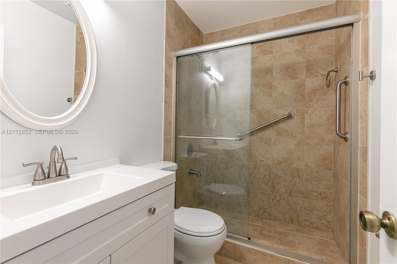 White vanity with integrated rectangular sink, oval wall-mounted mirror, and glass-enclosed shower with tiled surround and metal grab bars