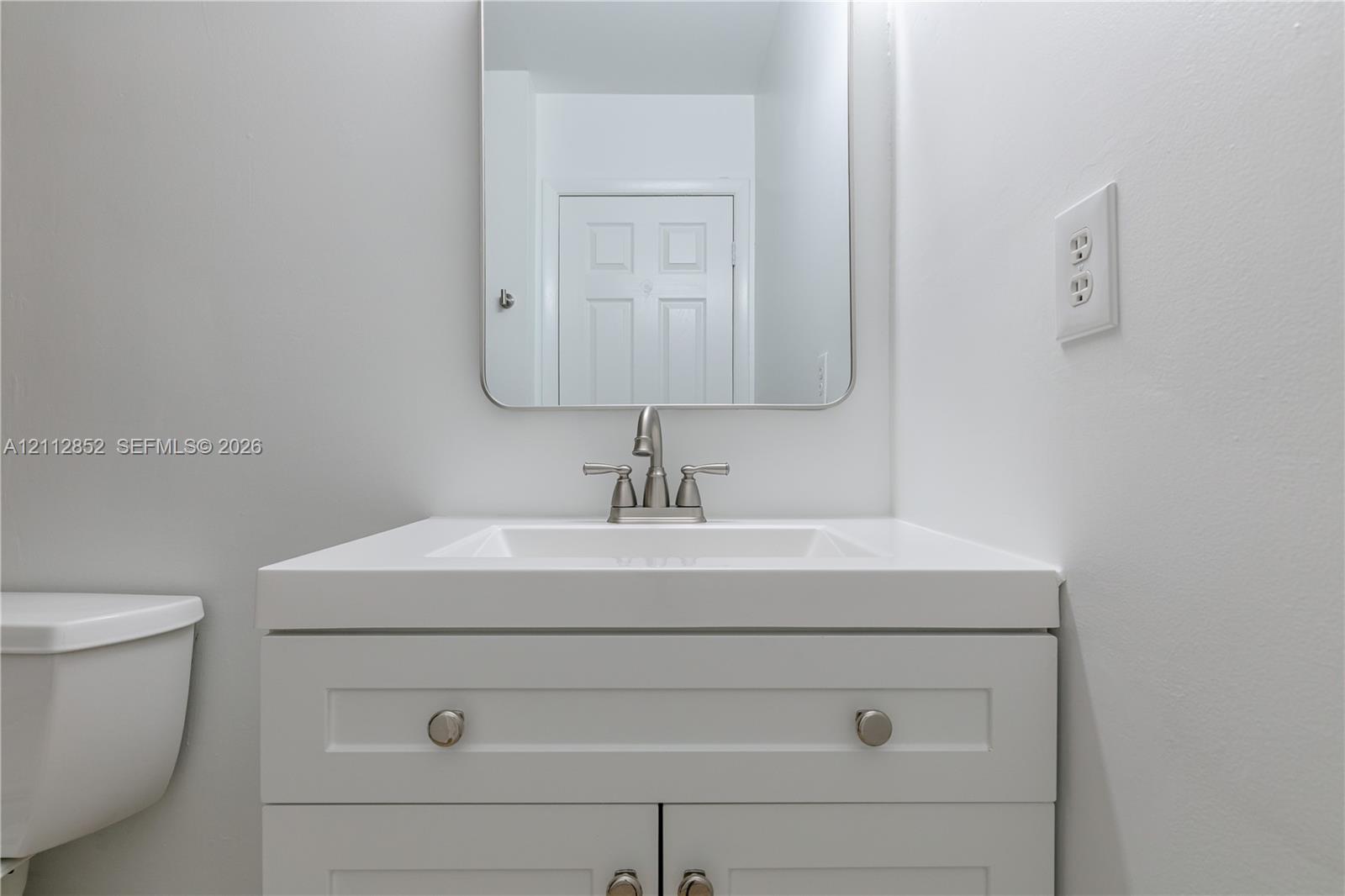 White vanity cabinet featuring a solid surface countertop with integrated basin, polished metal double-handle faucet, and rounded rectangular mirror