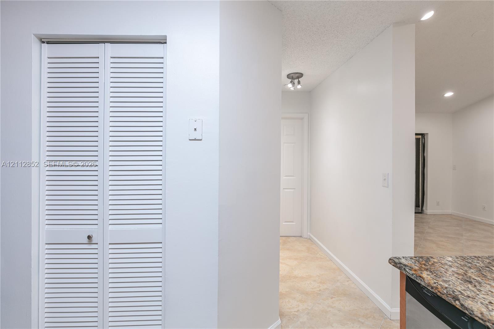 White louvered closet doors, neutral stone-finish tile flooring, multi-directional ceiling lighting, and a multi-toned stone countertop