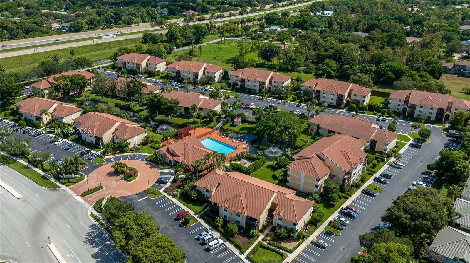 Aerial view of multi-unit residential complex featuring multi-story buildings with tile roofing, surface parking lots, a centralized outdoor swimming pool, and landscaped green space with water features
