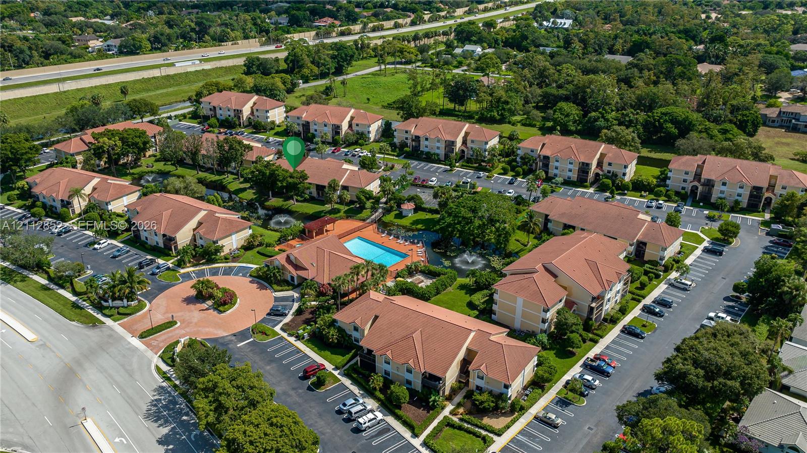 Aerial perspective of a multi-building residential community featuring tile roofing, a central swimming pool, landscaped grounds, and dedicated surface parking