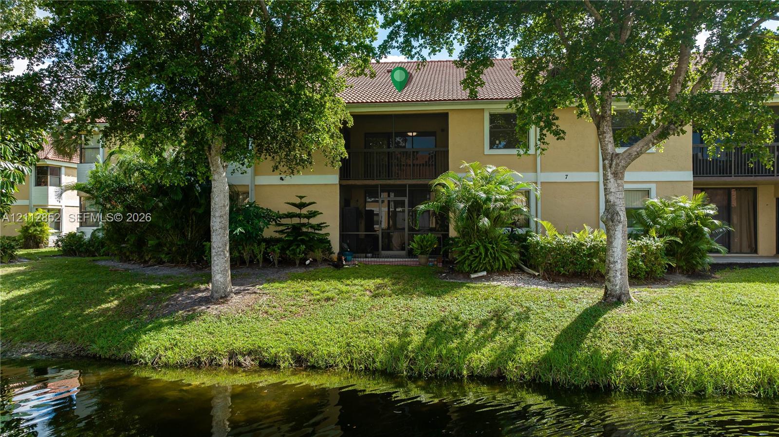 Two-story exterior featuring a tile roof, stucco finish, screened balcony, and water frontage with mature landscaping