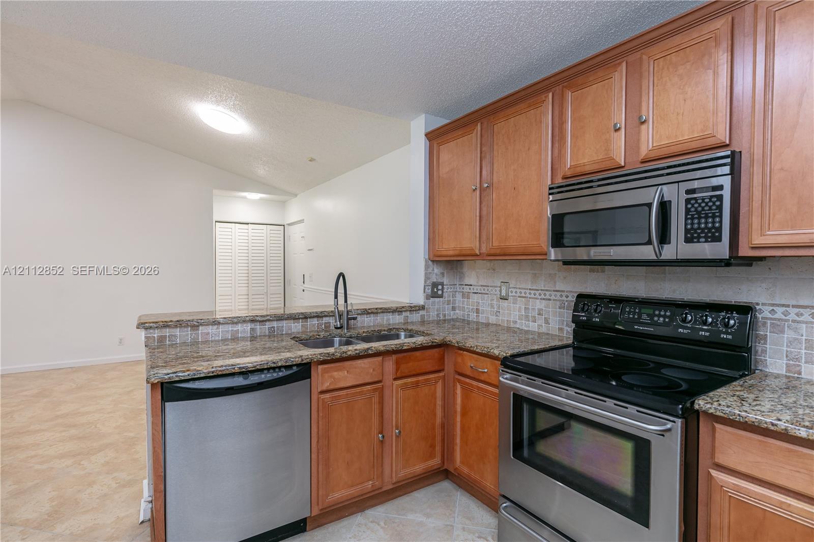 Kitchen featuring natural wood cabinetry, stone-surface countertops, stone tile backsplash, stainless steel appliances, and a double-basin sink with a high-arch pull-down faucet