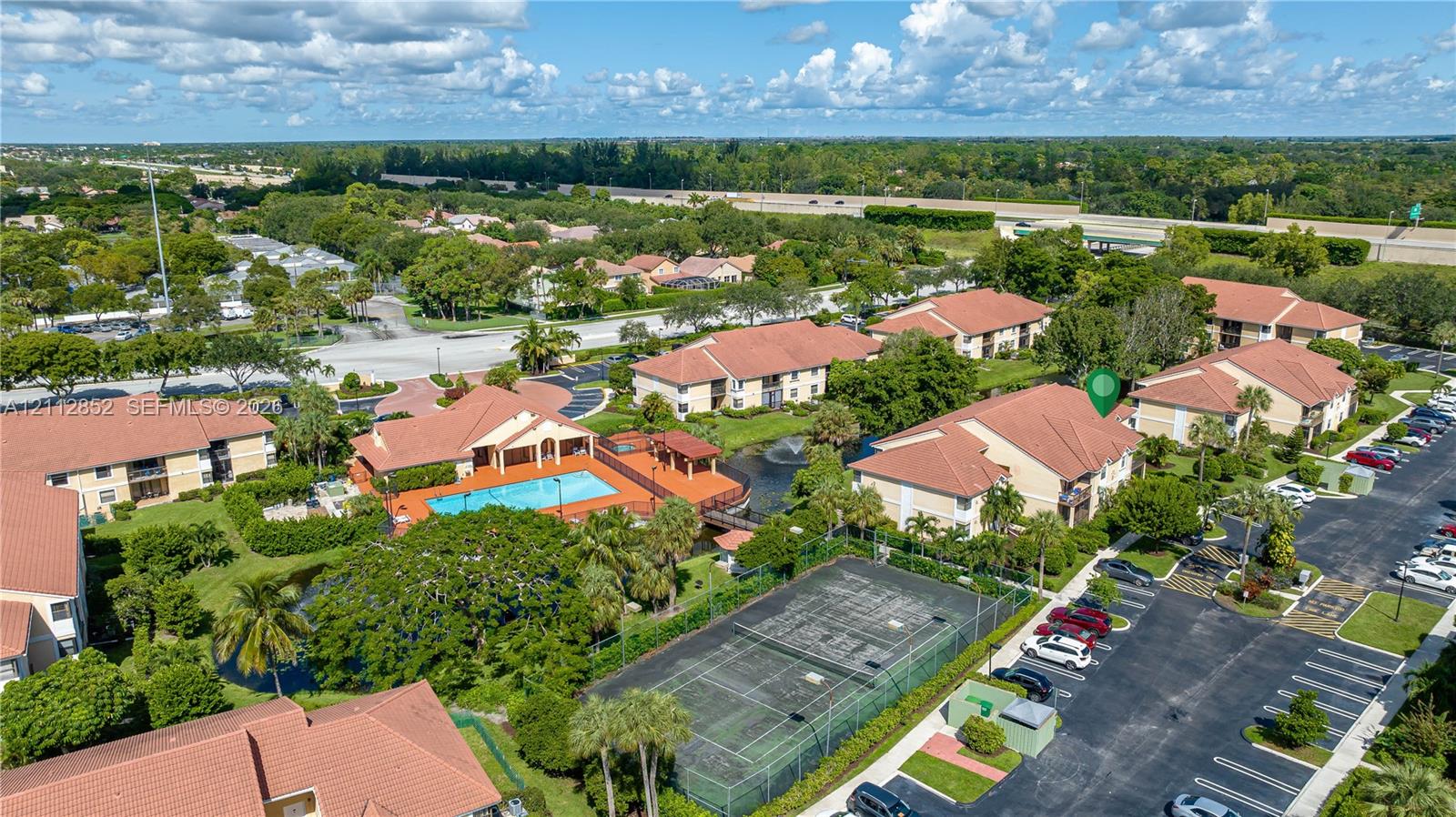 Aerial view of a residential community featuring a multi-story building, an outdoor swimming pool, a tennis court, and paved parking areas amidst landscaped grounds