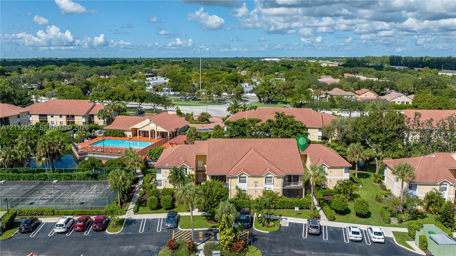 Aerial view of residential complex featuring a tile roof, paved parking area, landscaping, and community-accessible swimming pool and tennis court