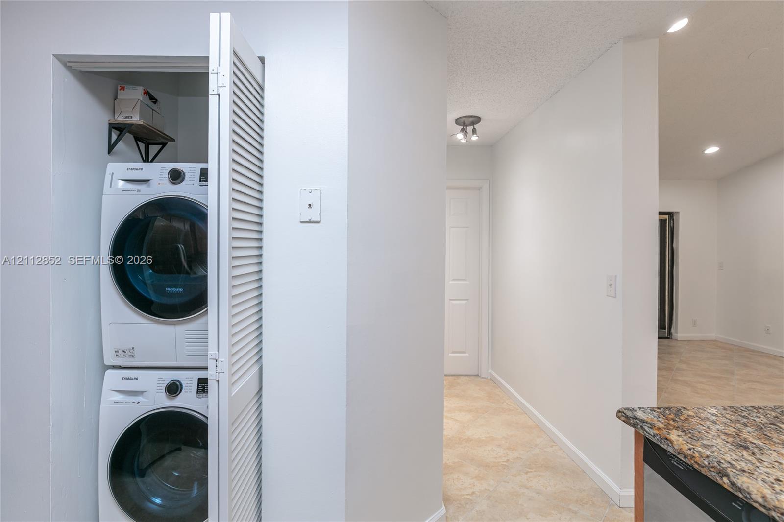 Stacked laundry unit featuring white appliances housed within a louvered-door closet, complemented by tile flooring and recessed lighting