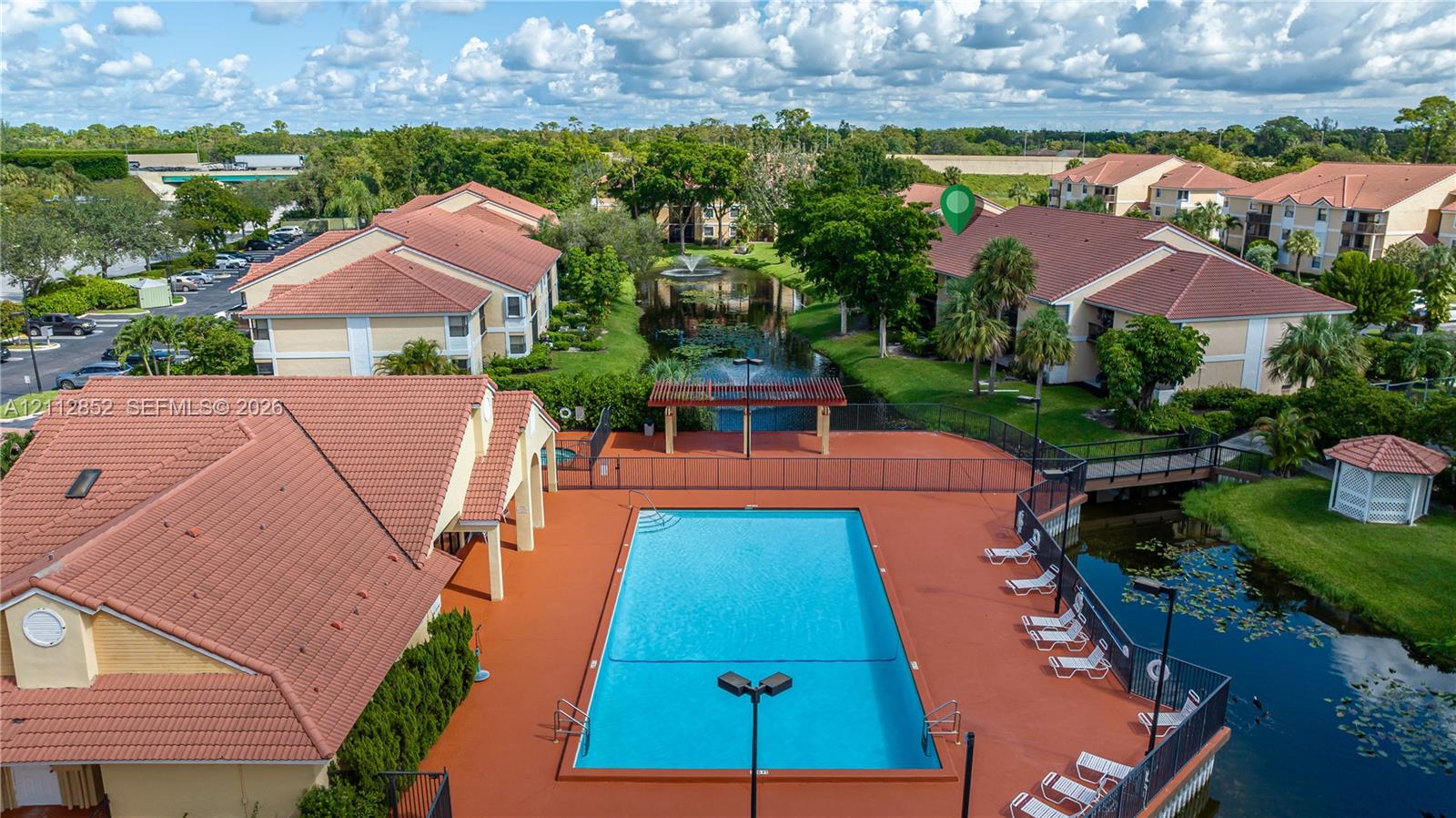 Community swimming pool with surrounding terrace, terracotta-style roof tiles, central water feature with fountain, covered pergola structure, and landscaped grounds