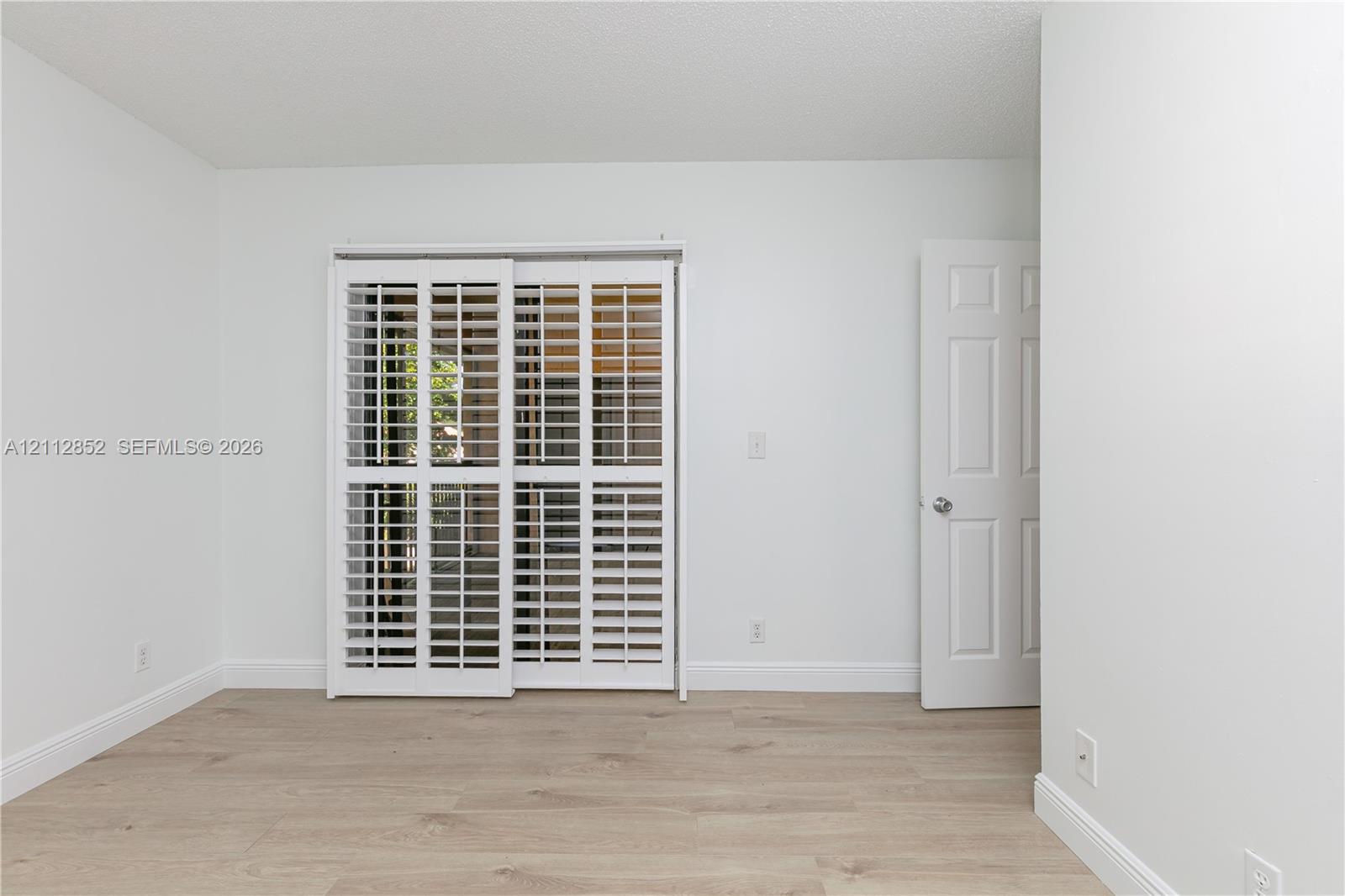 Interior space featuring light-toned wood-finish flooring, white walls, white plantation shutters on sliding doors, and a paneled swing door