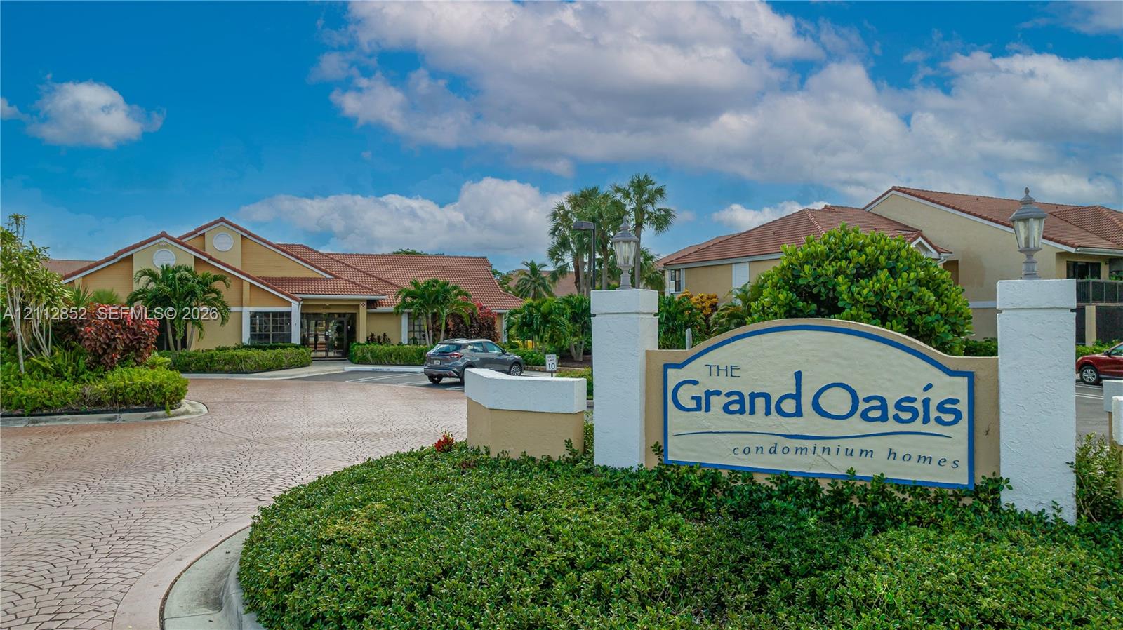 Community entrance featuring decorative stamped concrete paving, mature tropical landscaping, and a masonry monument sign with integrated pillar lighting