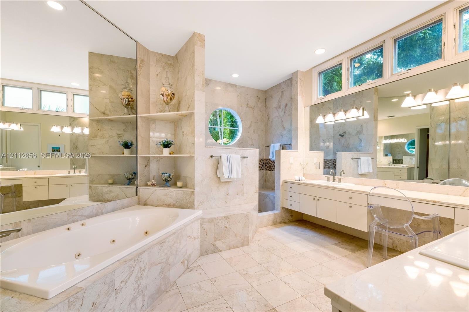 Spacious bathroom featuring stone-tiled surfaces, a built-in jetted tub, a circular accent window, dual vanities with integrated cabinetry, and extensive mirrored wall panels