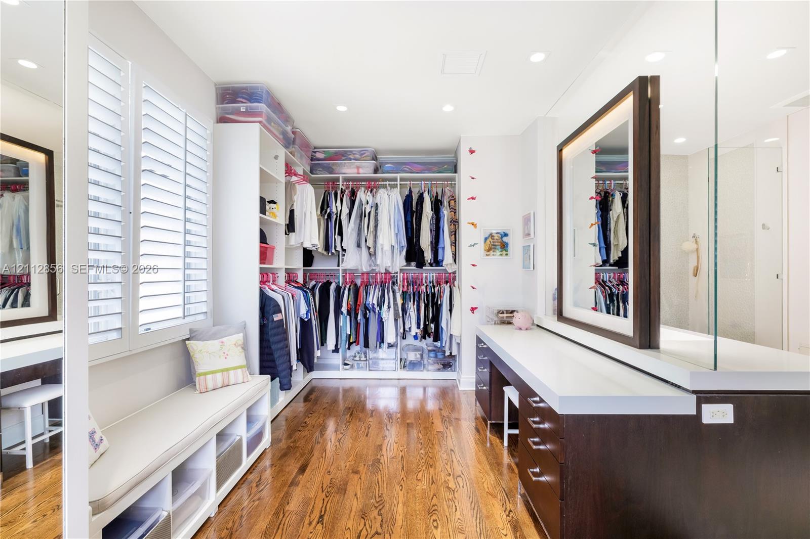 Custom dressing room featuring integrated floor-to-ceiling cabinetry, recessed lighting, wood-finish flooring, plantation shutters, and a built-in vanity with an illuminated mirror