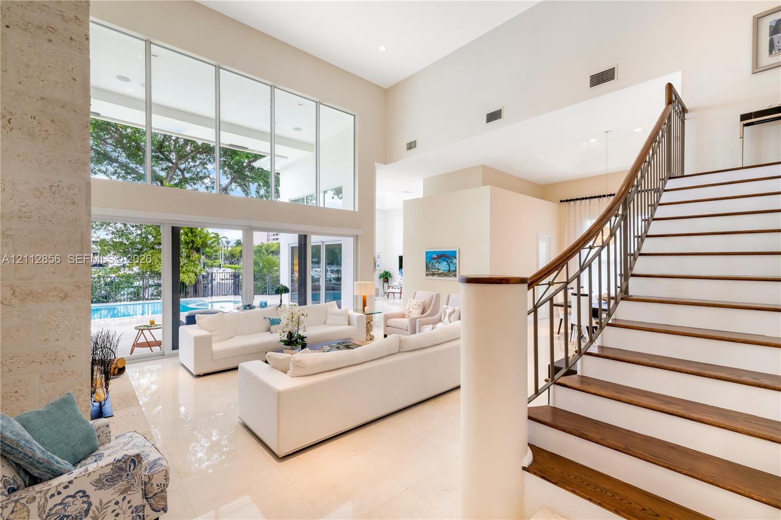 Double-height ceilings, floor-to-ceiling glass windows, staircase with wood-finish treads and metal railings, stone-faced interior column, and light-toned tile flooring