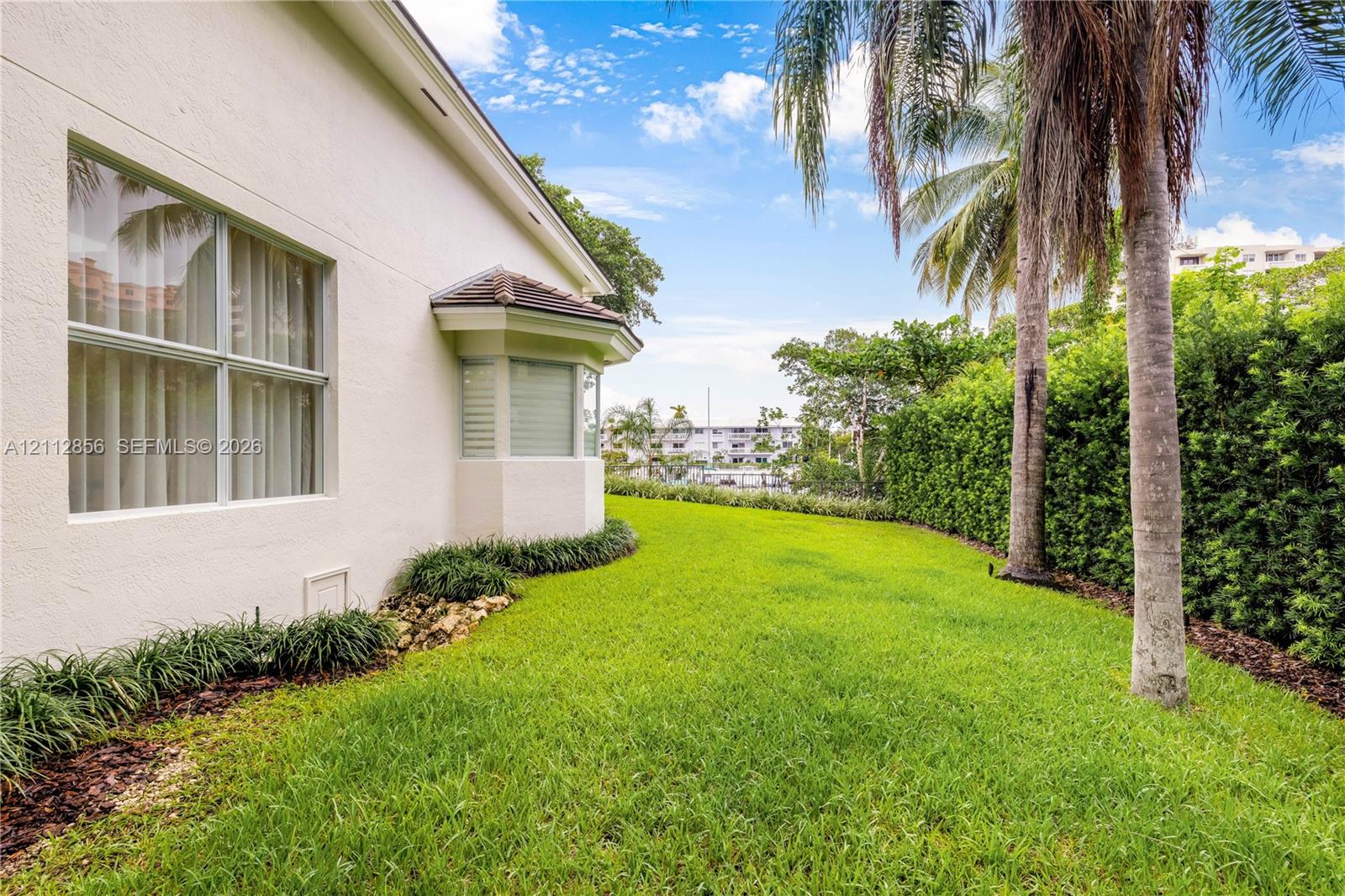 Stucco exterior featuring a bay window, expansive lawn, mature palm trees, and perimeter landscape hedging