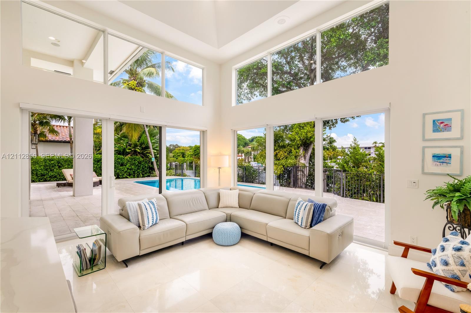 Double-height ceilings, floor-to-ceiling windows, polished stone flooring, and multiple sliding glass doors opening to a paved patio with a swimming pool