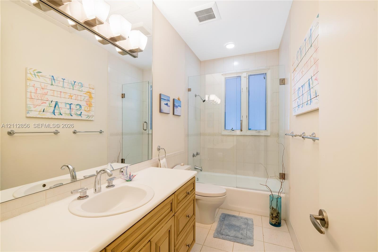 Bathroom featuring wood-finish cabinetry, an integrated sink with a chrome dual-handle faucet, a full-width vanity mirror, and a tub-shower combination enclosed by a frameless glass panel