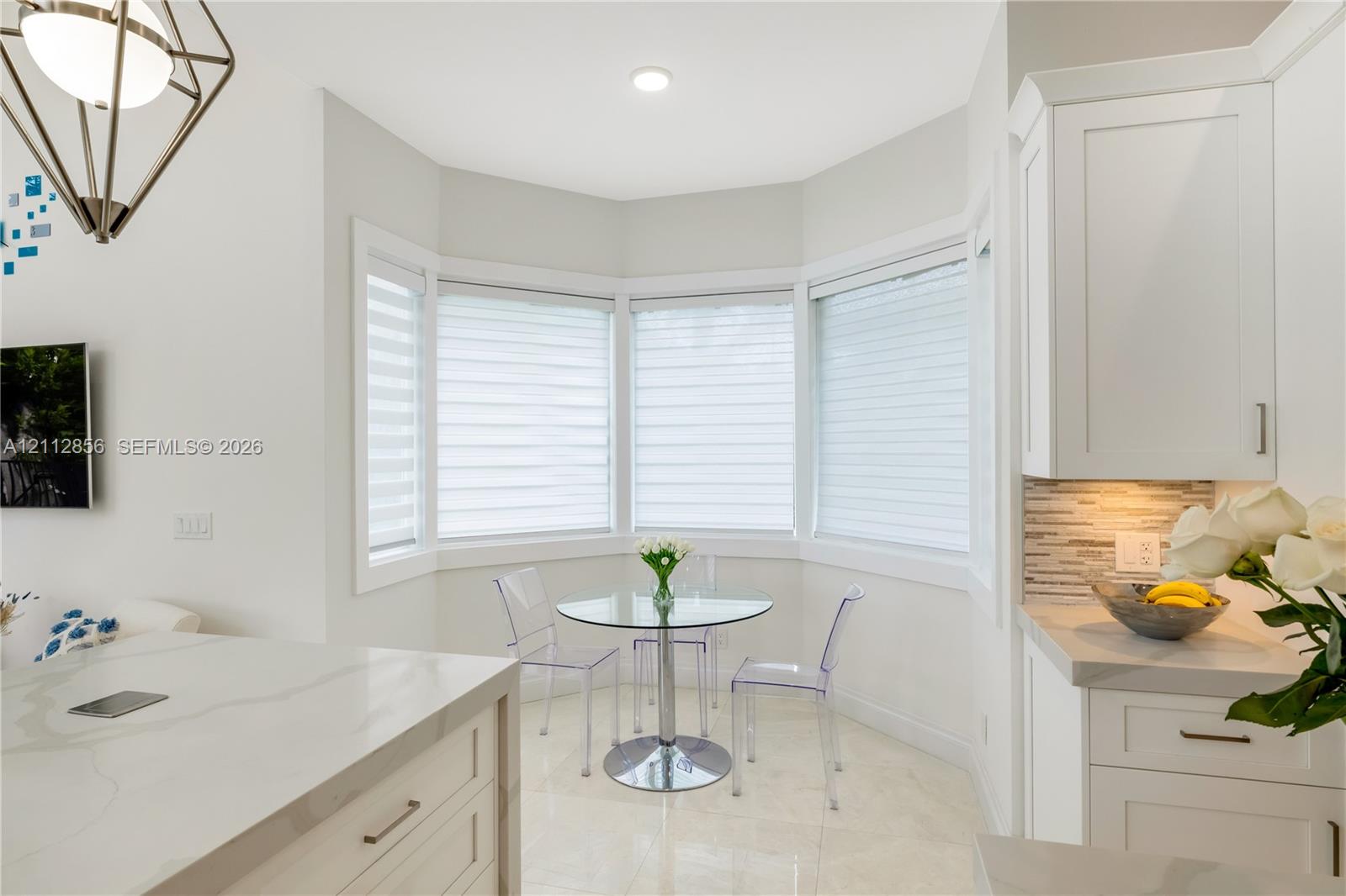 Kitchen nook featuring a bay window, white shaker-style cabinetry, stone-finish countertops, geometric metal pendant lighting, and neutral tile flooring