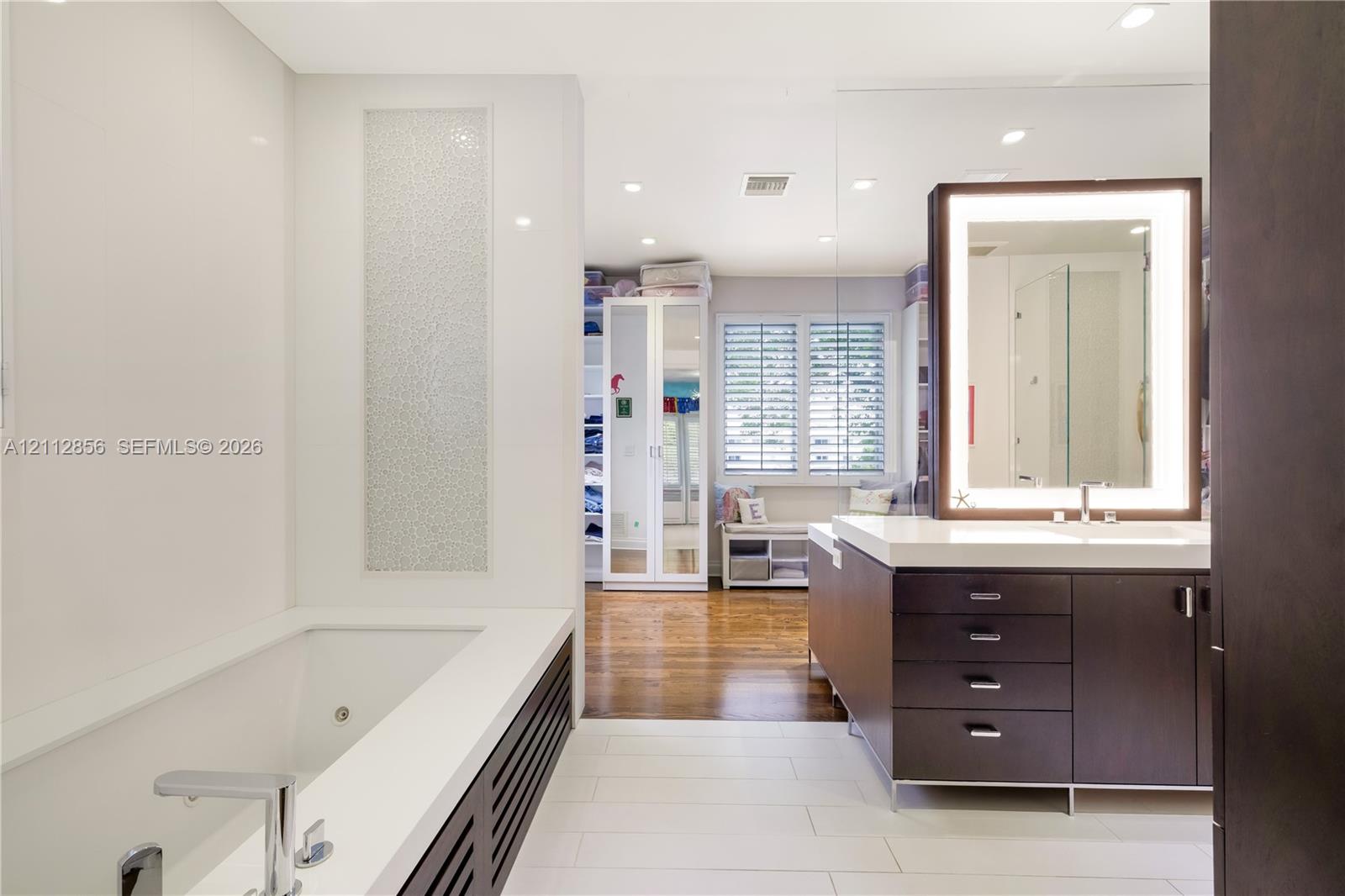Contemporary bathroom featuring a deep-soaking tub with a tiled surround, custom dark-finish vanity cabinetry with a stone countertop, an integrated LED-lighted mirror, and large-format porcelain floor tile