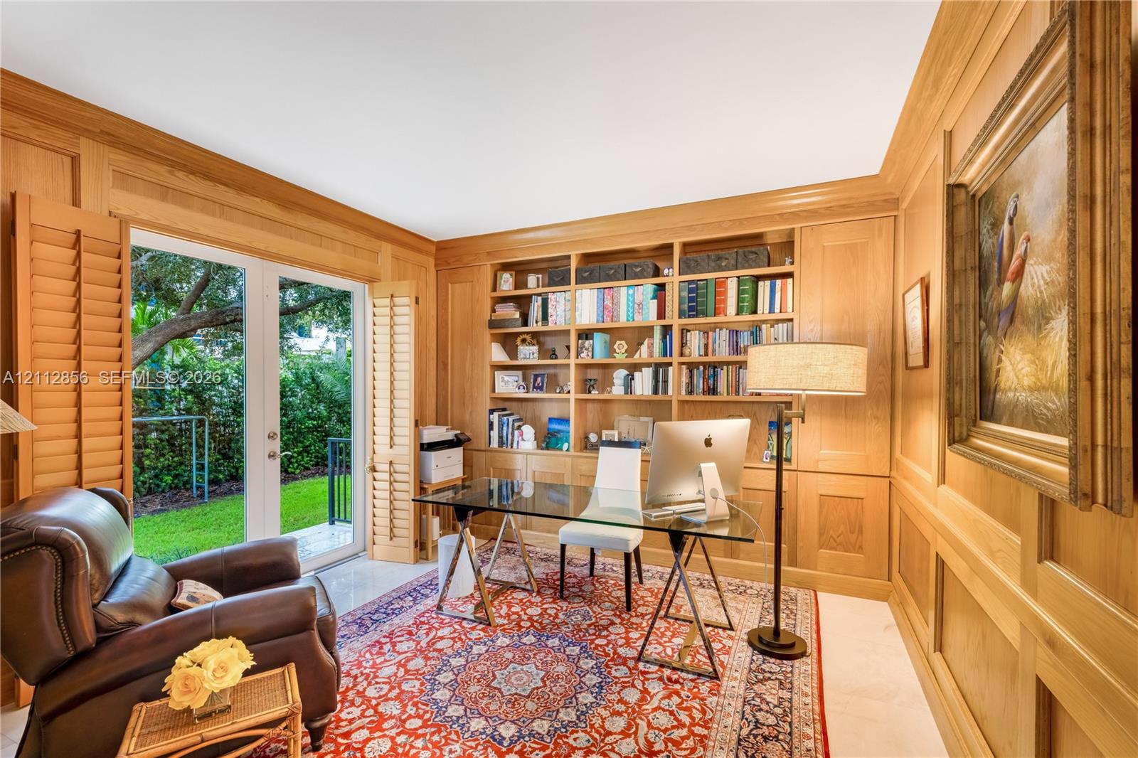 Wood-paneled room featuring custom floor-to-ceiling built-in bookshelves, light-finish tile flooring, and glass French doors with integrated louvered shutters