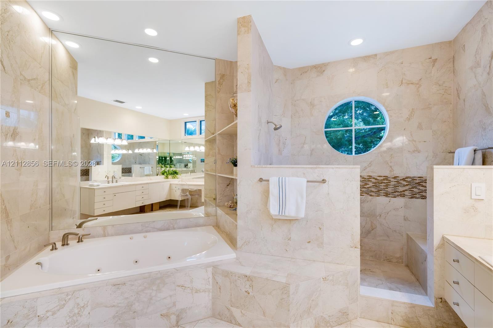 Stone-tiled bathroom featuring a jetted corner tub, walk-in shower with circular window, expansive wall-to-wall mirror, and built-in shelving