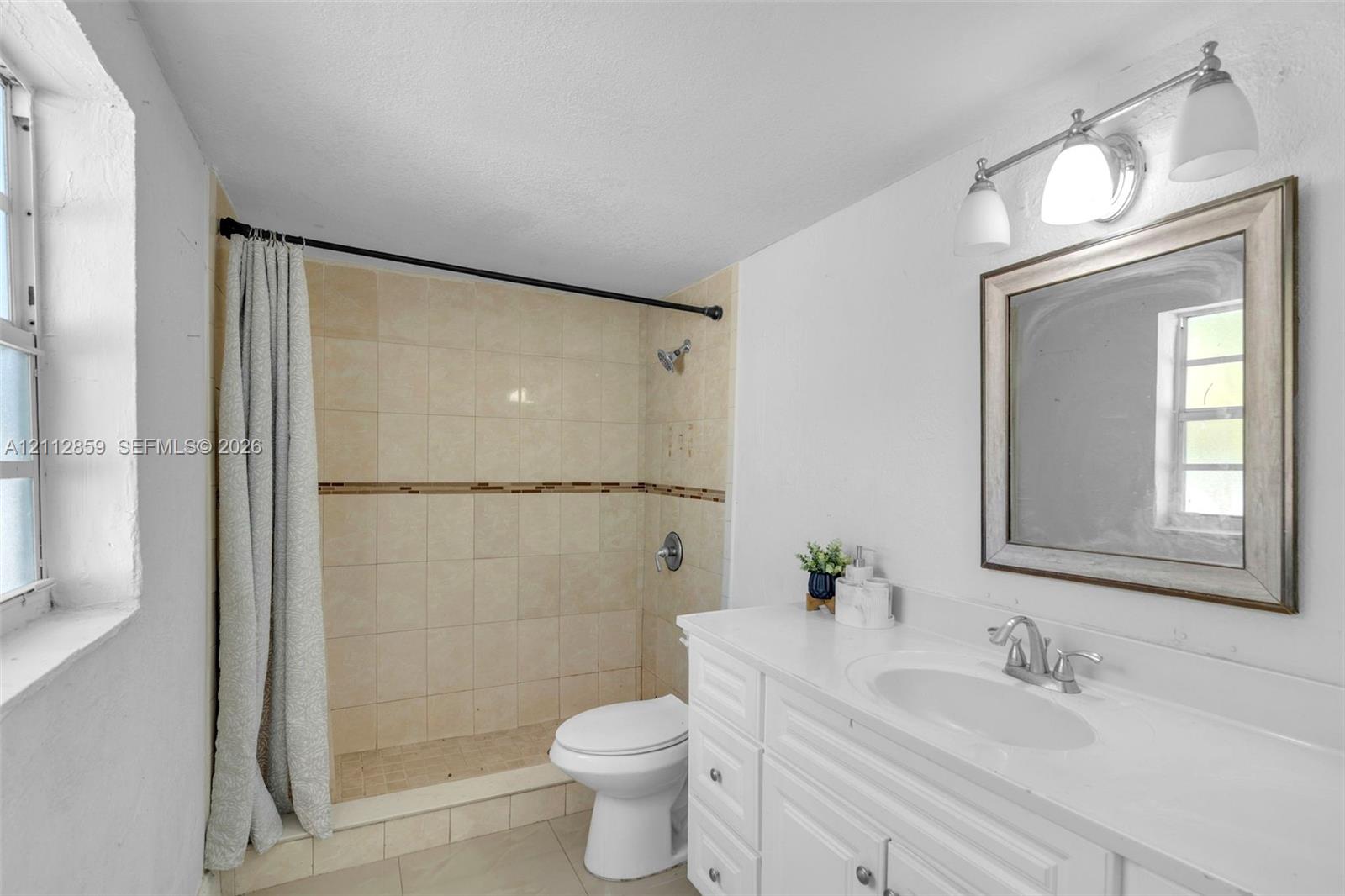 Bathroom featuring a white vanity with an integrated sink, a rectangular framed mirror, a triple-light fixture, and a tiled shower stall with a decorative accent border
