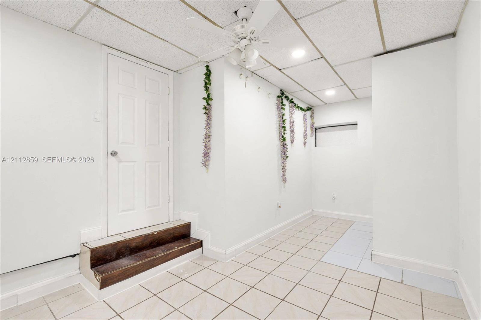Tiled flooring, drop ceiling with recessed lighting, integrated ceiling fan, and wood-finish step leading to a six-panel door