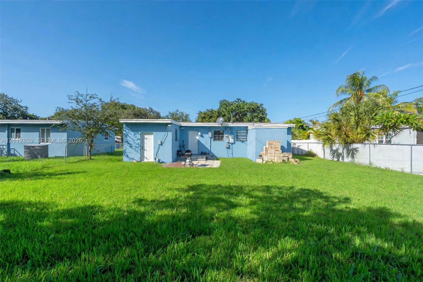 Expansive backyard featuring a large grass lawn, perimeter chain-link fencing, mature trees, and a blue stucco exterior