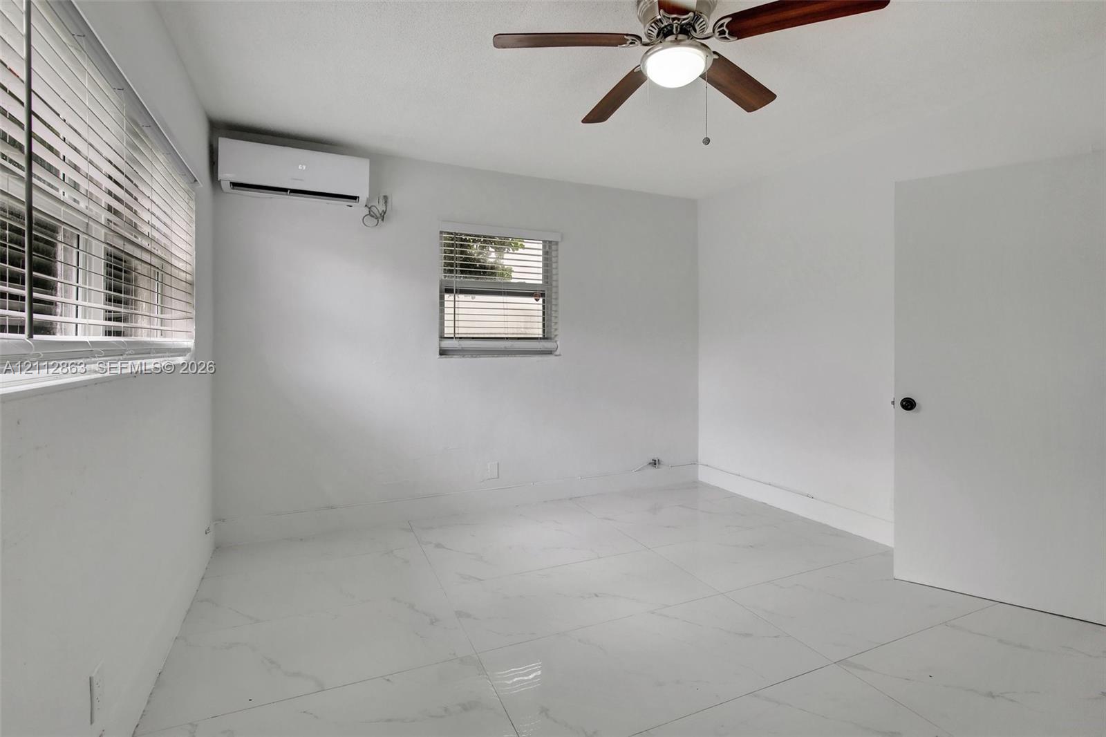 Large-format polished tile flooring, wall-mounted ductless air conditioning unit, ceiling fan with integrated lighting, and horizontal window blinds