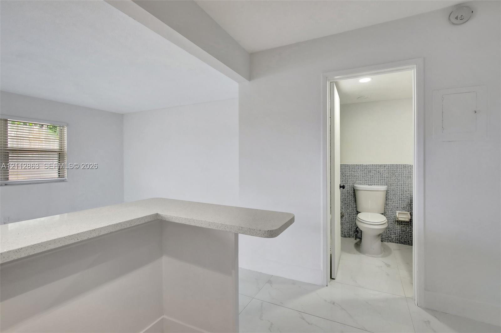 Neutral-toned kitchen counter with a matte finish, white porcelain tile flooring, and a bathroom featuring mosaic tile wall accents