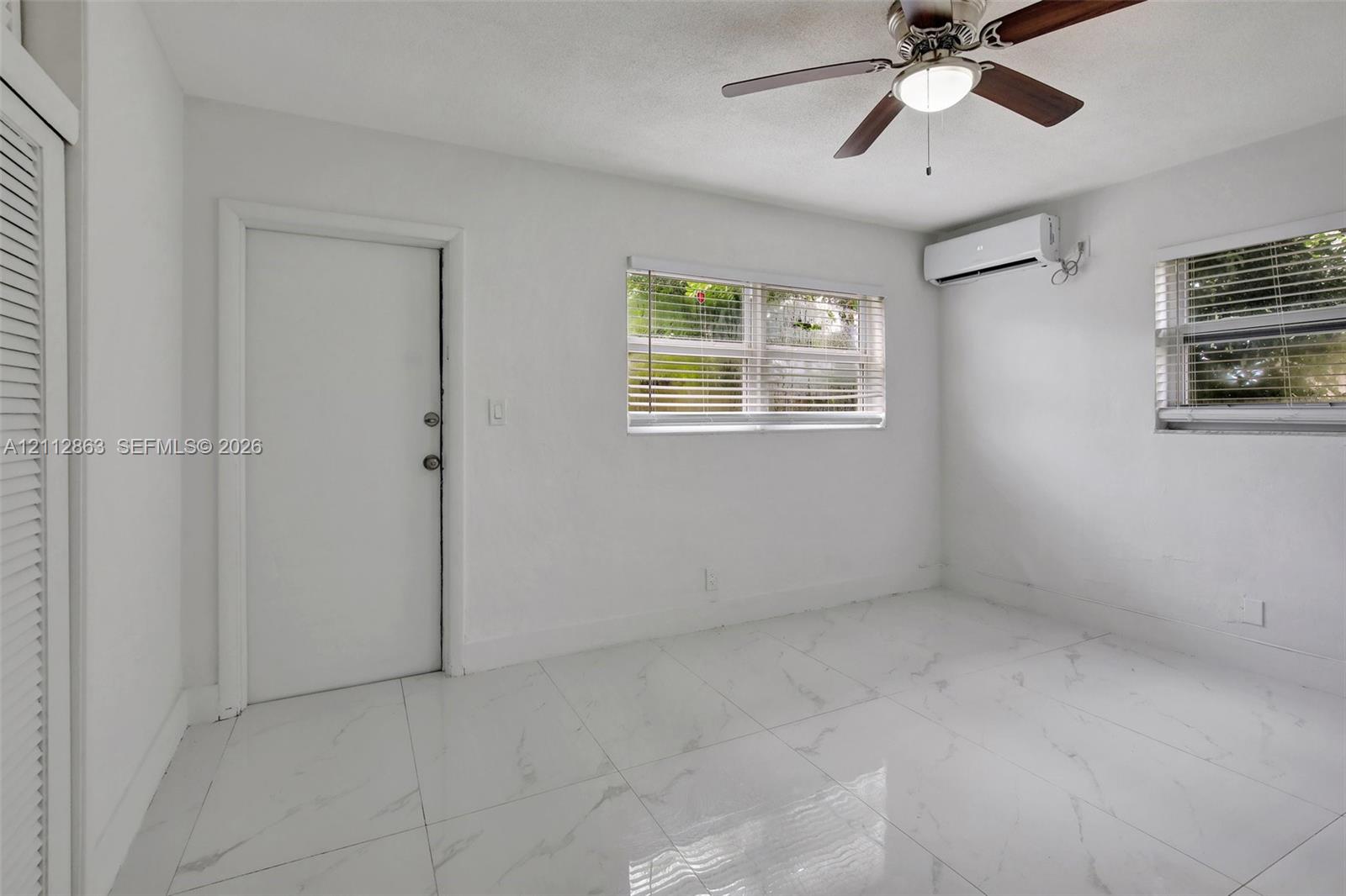 Room featuring large-format white tile flooring, wall-mounted ductless air conditioning unit, ceiling fan with integrated lighting, and louvered closet doors
