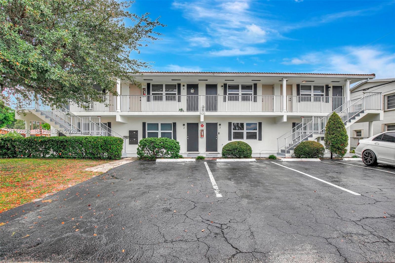Two-story exterior featuring white stucco siding, dual-level railed balconies, paved surface parking, and manicured shrubbery