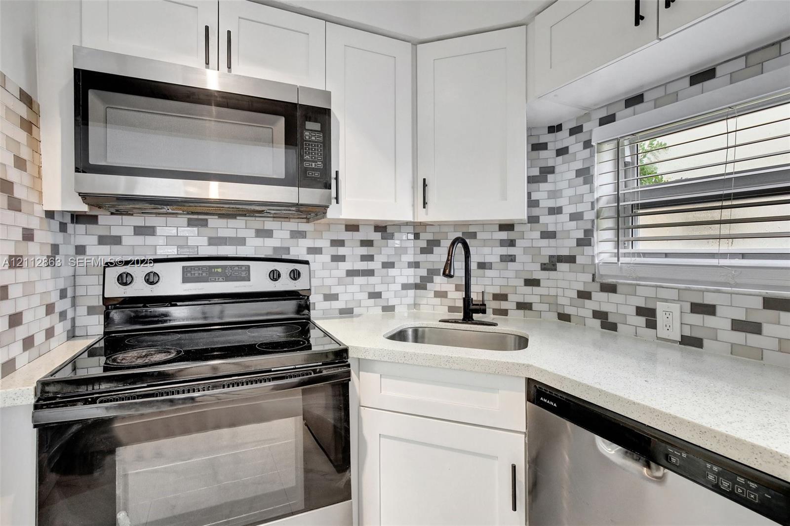 Kitchen featuring white shaker-style cabinetry, a multi-tonal rectangular tile backsplash, stainless steel appliances, light-colored countertops, and a matte black gooseneck faucet