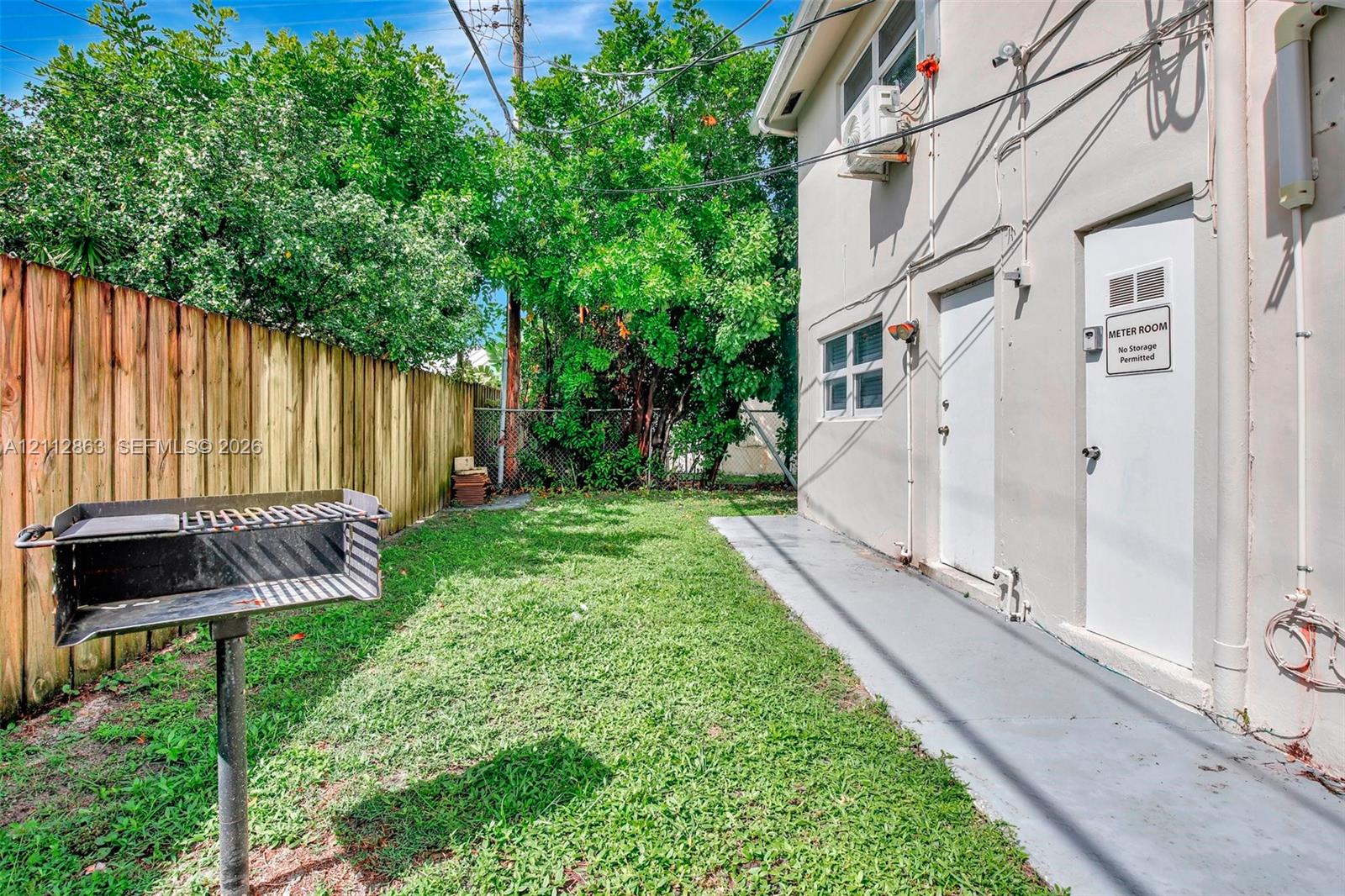 Exterior side yard featuring a wood-panel fence, a stationary metal grill, and a concrete walkway
