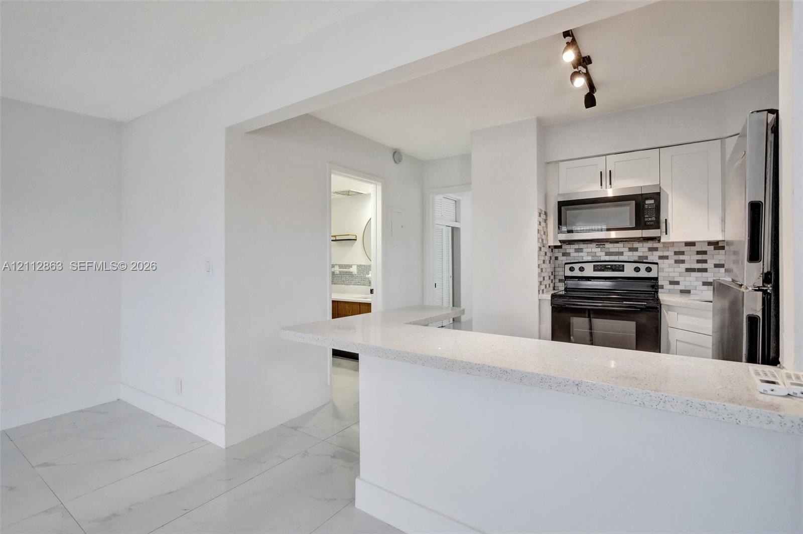Kitchen featuring white shaker-style cabinetry, a quartz countertop breakfast bar, a stone-tiled backsplash, stainless steel appliances, and large-format porcelain floor tiling