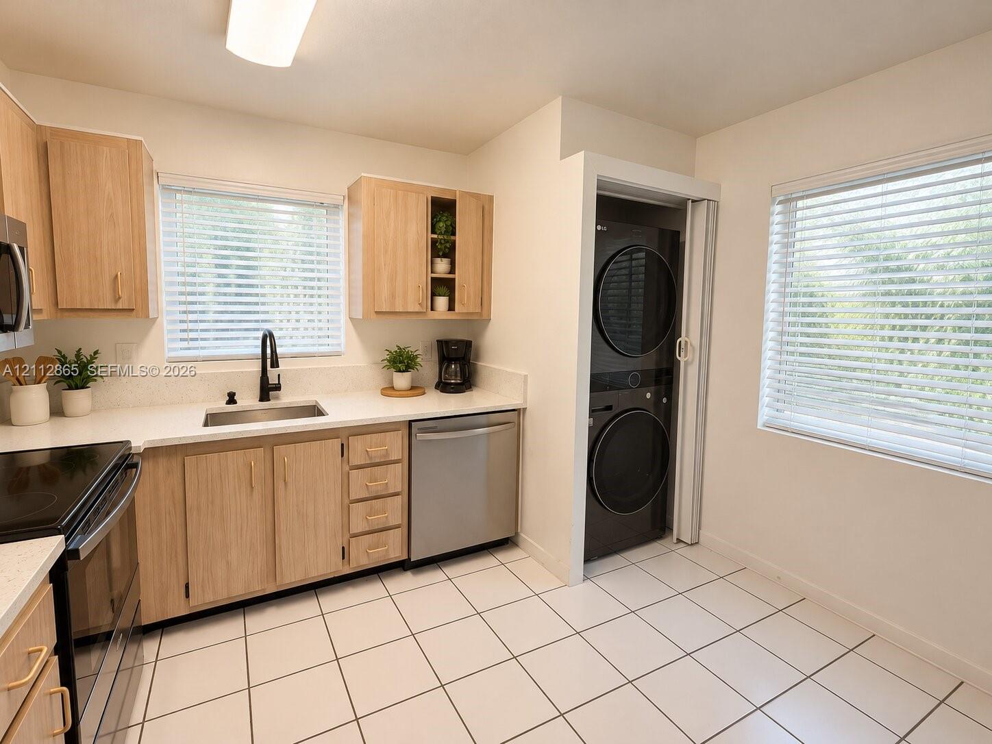 Kitchen featuring light wood-finish cabinetry, white solid-surface countertops, stainless steel undermount sink with matte black faucet, ceramic tile flooring, and a built-in laundry closet