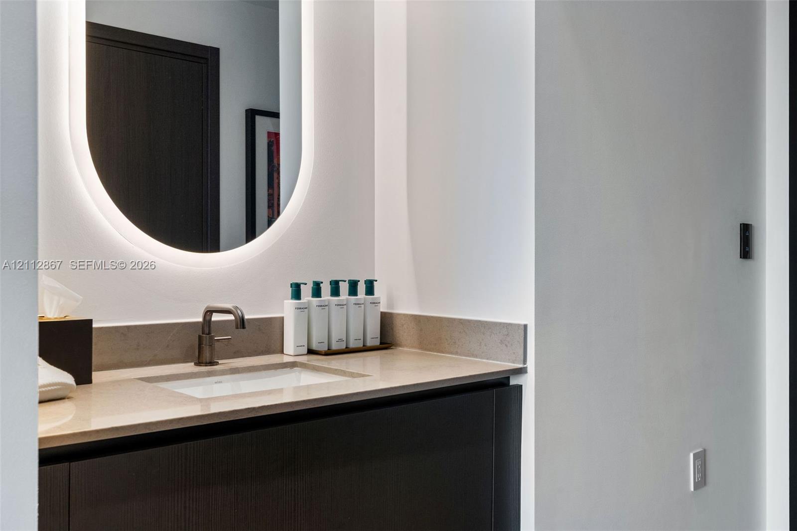 Dark-finish cabinetry with a neutral stone countertop, integrated rectangular sink, wall-mounted metal faucet, and backlit oval mirror Dark-finish cabinetry with a neutral stone countertop, integrated rectangular sink, wall-mounted metal faucet, and backlit oval mirror