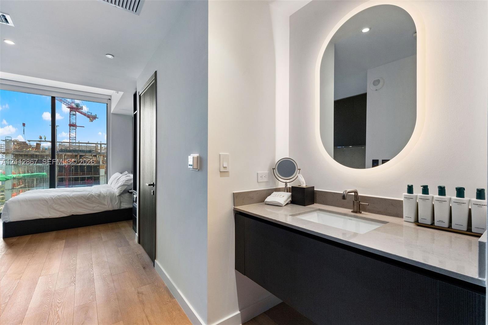 Modern floating vanity with a neutral-tone countertop, undermount sink, and metallic faucet, complemented by a backlit oval wall mirror and wood-finish flooring Modern floating vanity with a neutral-tone countertop, undermount sink, and metallic faucet, complemented by a backlit oval wall mirror and wood-finish flooring