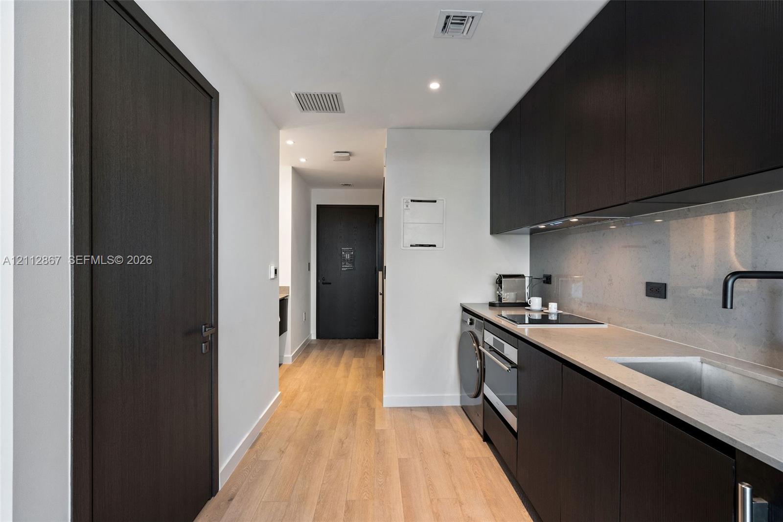 Kitchenette featuring dark-finish cabinetry, solid-surface countertops, integrated cooktop and oven, under-mount sink with matte black faucet, and wood-finish flooring Kitchenette featuring dark-finish cabinetry, solid-surface countertops, integrated cooktop and oven, under-mount sink with matte black faucet, and wood-finish flooring