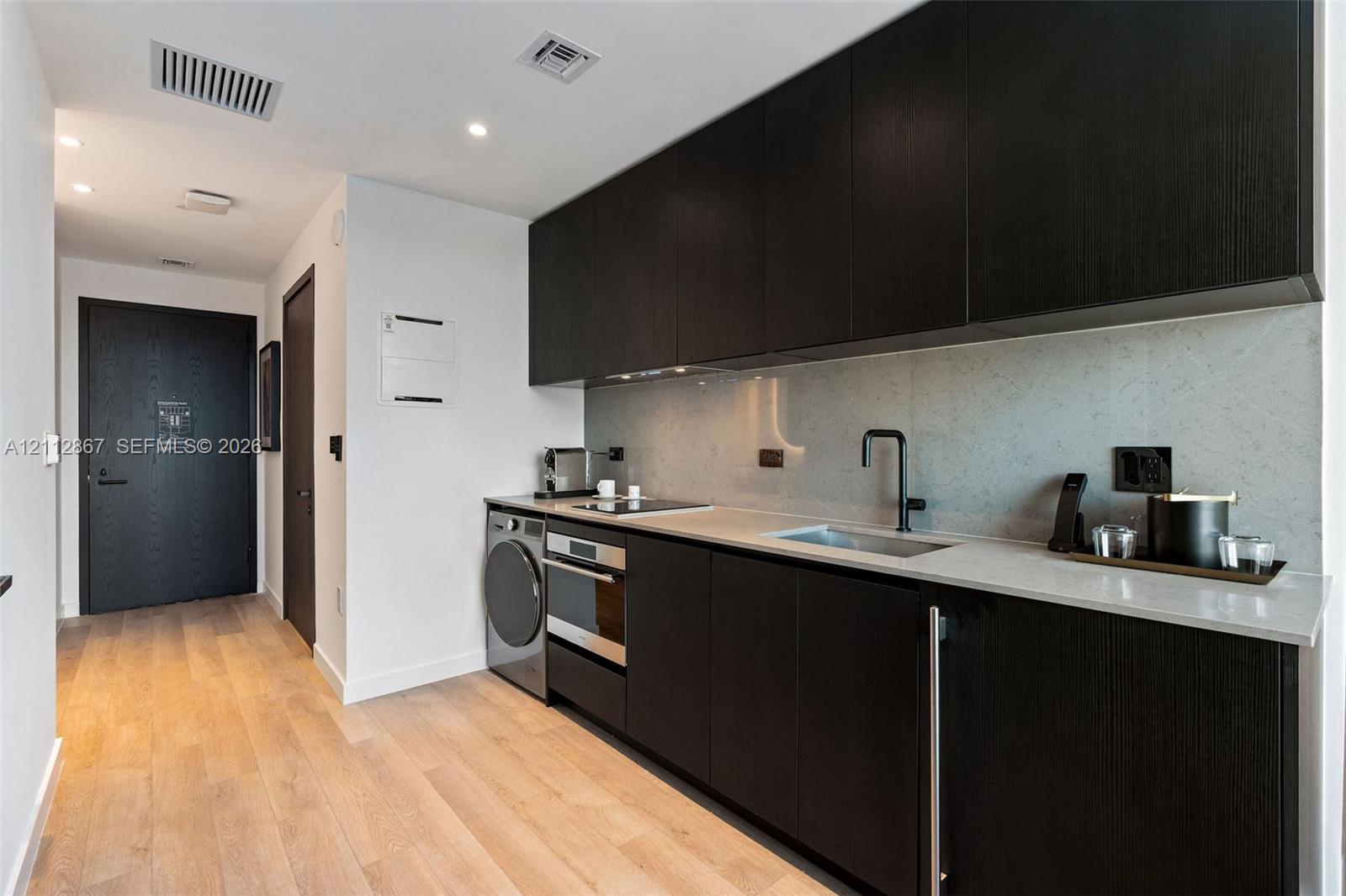 Kitchen featuring dark wood-finish cabinetry, solid surface countertops, a full-height stone backsplash, integrated stainless steel appliances, and wood-finish flooring Kitchen featuring dark wood-finish cabinetry, solid surface countertops, a full-height stone backsplash, integrated stainless steel appliances, and wood-finish flooring